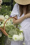 Person i vit klänning håller en korg med vita sommarblommor i en grön trädgård.