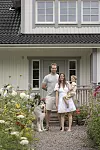 Familj med barn och hund står på grusgång framför vit villa omgiven av blomrabatter.