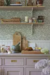 Lantligt kök med träbänk, mortel, kakform, snittblommor och hyllor mot blommig tapet.
