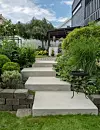 Betongtrappa leder genom frodig trädgård upp till trädäck vid ett modernt hus.