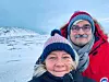 Två personer i vinterkläder tar en selfie i ett snötäckt landskap med berg i bakgrunden.