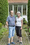 Äldre par står på stenlagd trädgårdsgång framför ett hus med glasveranda.