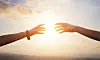 Två händer når mot varandra framför en stark sol vid horisonten.