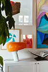 Orange bordslampa på vit skänk framför färgglada tavlor på väggen.