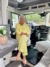 Äldre kvinna i gul dräkt sitter barfota inne i en husbil parkerad vid tallskog.