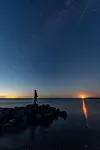 Person står på klippor vid havet under klar stjärnhimmel med måne vid horisonten.
