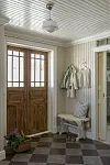 Ljus hall med dubbla trädörrar, randig vägg, bänk, krokar med ytterkläder och rutigt stengolv.