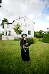 Äldre kvinna med blombukett står på gräsmatta framför stort vitputsat hus.