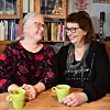 Två leende kvinnor sitter vid ett köksbord med gröna kaffekoppar framför en bokhylla.