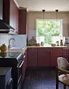 Kök med bordeauxfärgade skåp, träbänkskiva och stort fönster mot grönskande trädgård.