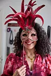 Leende kvinna i blommig röd blus håller upp en röd fjädermask framför ansiktet inomhus.