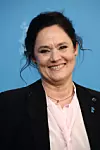 Leende kvinna i kavaj och ljusrosa blus framför blå bakgrund.
