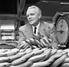 Kostymklädd man står bakom ett stort parti bananer i en lagerlokal.
