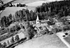 Flygfoto över lantlig kyrka med kyrkogård omgiven av skog och åkrar.