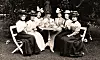 Svartvitt foto av kvinnor i långa kjolar och hattar runt ett kaffebord utomhus.