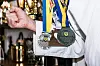 Arm i vit dräkt med flera medaljer och blågula band framför pokaler i bakgrunden.