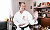 Äldre man i karate-gi demonstrerar ställning i vardagsrum med pokaler bakom sig.