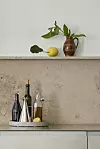 Stilleben i kök med flaskor, karaff, citron och brun kanna med gröna blad mot stenvägg.