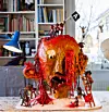 Stort färgstarkt skulpturhuvud med små figurer som klättrar och gräver i en ateljé fylld av böcker.