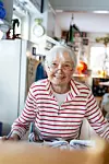 Leende äldre kvinna i rödvitrandig tröja sitter och tecknar i en ljus ateljé.