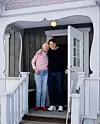 Äldre kvinna och man står omfamnande i en ytterdörr till ett trähus.