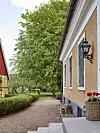 Gult stenhus med grusgång, häckar och stort grönt träd vid entrén.
