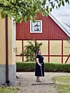 Kvinna i svart klänning står med kratta på gårdsplan framför gul och röd gårdsbyggnad.