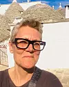 Person med svarta glasögon framför vit stenbyggnad med konformade tak i starkt solsken.