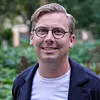 Person med glasögon och kavaj ler mot kameran utomhus med grönska i bakgrunden.