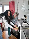 Yngre kvinna sätter in en form i ugnen bredvid leende äldre kvinna i kök.