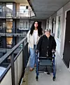 Ung kvinna går bredvid äldre kvinna med rullator på loftgång utanför lägenhet.