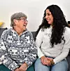 Äldre kvinna och yngre kvinna sitter i soffa och ler mot varandra.