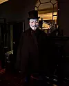 Person i Guy Fawkes-mask och hög hatt står i ett mörkt, elegant rum.
