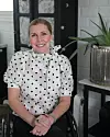 Leende kvinna i vit prickig blus sitter framför mörka skåp och en grön växt.