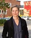 Person med kort hår står utomhus framför tegelhus och parkerade bilar.