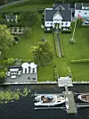 Flygfoto över vit villa med stor trädgård, soldäck och båtar vid brygga i en älv.