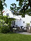 Vitmålat trähus med stenlagd uteplats, rottingmöbler och lummig trädgård.