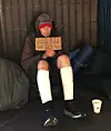 Hemlös person sitter mot en vägg med kartongskylt och bandagerade ben.