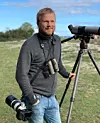 Person med kikare och tubkikare på stativ står utomhus på en öppen naturplats.