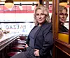 Person i kavaj lutar mot spegelvägg i bar med röda barstolar och stora fönster.