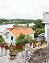 Altan med dukat bord, krukväxter och utsikt över vita trähus och sjö.