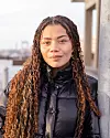 Person i svart dunjacka med långt lockigt hår står vid kaj med stad i bakgrunden.
