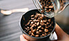 Rostade kaffebönor hälls från glasburk ner i kaffekvarn