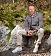 Man sitter avslappnat på stenar utomhus framför en sluttning med gräs och buskar.