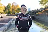 Ung person i stickad tröja står vid kanal omgivet av höstlöv och stadsmiljö.