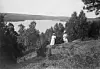 Två kvinnor i ljusa klänningar står på en höjd med utsikt över en sjö omgiven av skog.