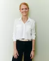 Dorthe Snejbjerg står op ad lys væg iført hvid skjorte og mørke bukser og smiler til kameraet.