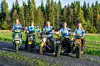 Fem personer står med varsin moped på en grusväg framför en skog i kvällssol.