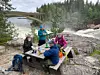 Fyra personer fikar vid ett träbord på klippor intill en fors och en bro i skogsmiljö.
