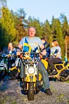 Person på gul moped med svenska flaggor, omgiven av familj på mopeder i kvällssol.